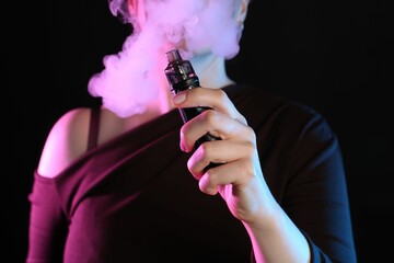 Woman using electronic smoking device in neon lights on black background, closeup