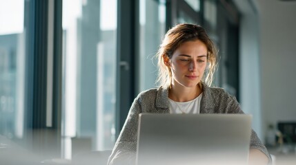 Professional young woman working on laptop in modern office with large windows.