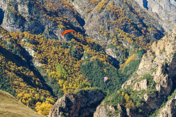 Paragliders soar over rugged mountain landscape adorned with autumn foliage. Fly Chegem paragliding site, El-Tyubu, Russia - October 7, 2024