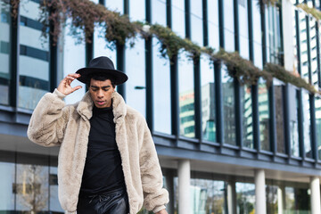 Unemotional adult ethnic male in trendy apparel hat and fur jacket walking on the street near glass...