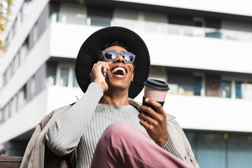 Stylish ethnic man talking with smartphone and coffee to go outdoors. Spanish American male in...