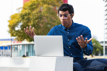Indian man sitting in city street looking at laptop typing browsing working loses becoming...