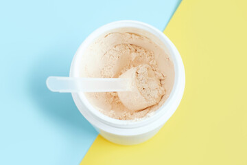 Protein powder in scoop and plastic jar on blue and yellow background.