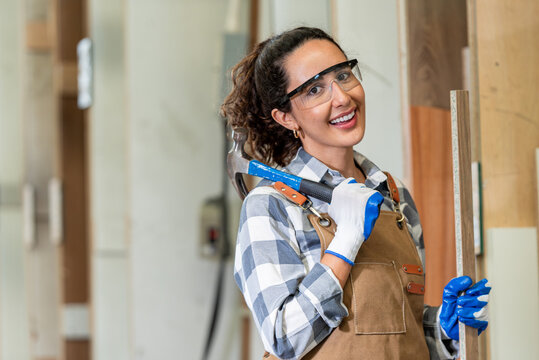 One Strong confident young aged women carpenter standing look at camera in workshop. Latin female carpenter entrepreneur working with wood plank furniture tool in workbench shop carpentry - Powered by Adobe