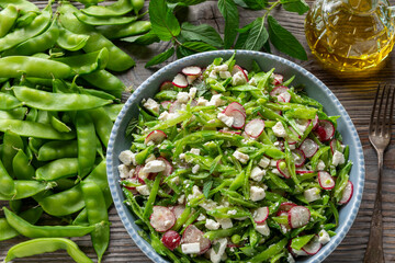 Sugar snap pea and radish salad with torn mint leaves and aged ricotta cheese. Pods of green sugar snap peas, sprig of mint, and bottle of olive oil on the side. Rustic wooden table.