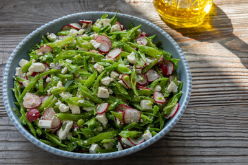 Sugar snap pea and radish salad with torn mint leaves and aged ricotta cheese. A bottle of olive oil behind. Rustic wooden table.