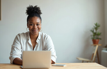 Smiling African American woman at home workplace. Woman sits desk looking at camera. Black female using laptop. Businesswoman works at home minimal interior. Happy, confident, positive attitude.