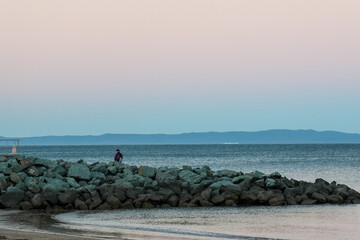 Solitary figure sitting on rocky breakwater overlooking calm ocean at dawn with soft pastel sky and...
