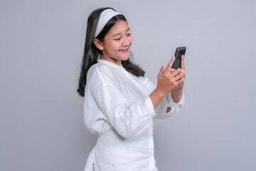 Cheerful young Asian woman in a white shirt and headband smiling while using a smartphone, looking at the screen with joy and excitement, isolated on a plain gray background.