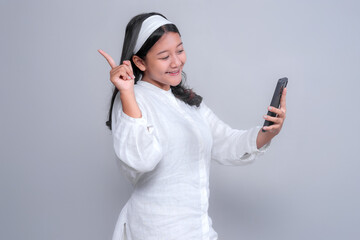 Joyful young Asian woman in a white shirt and headband smiling while holding a smartphone and pointing upward, expressing excitement or new idea, isolated on a clean gray background.