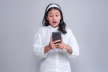 Surprised young Asian woman wearing a white shirt and headband looking at her smartphone with wide eyes and open mouth, reacting to unexpected news or shocking information.