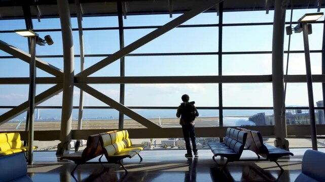 Traveler man with backpack walking to look at runway and planes through large window in airport terminal with bright sunlight. Waiting for flight during vacation. Kansai international airport.