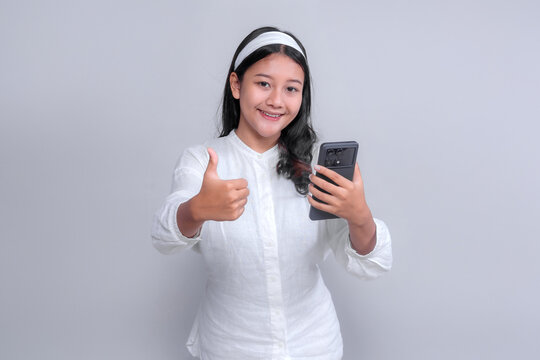 Asian woman wearing white shirt and headband smiling confidently while holding smartphone and showing thumbs up gesture, isolated on gray background, technology concept.
