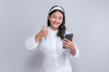 Asian woman wearing white shirt and headband smiling confidently while holding smartphone and showing thumbs up gesture, isolated on gray background, technology concept.