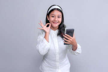 Asian woman with long black hair wearing a white shirt and headband, smiling while holding a smartphone in one hand and making an OK hand gesture with the other.