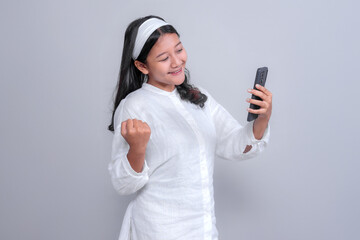 Asian woman wearing a white shirt and headband, holding smartphone while smiling and raising her fist in a happy, victorious pose, isolated on grey background, studio shot.