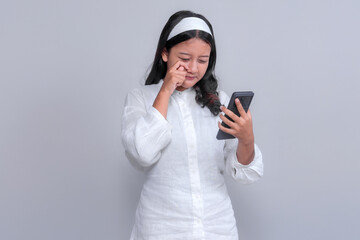 Asian woman wearing a white shirt and headband looking at her smartphone with a sad facial expression, wiping tears, isolated on grey background, studio portrait.