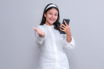 Asian woman wearing a white shirt and headband holding a smartphone and gesturing with her hand as if inviting or presenting, smiling, isolated on grey background.