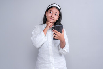 Thoughtful young Asian woman in a white shirt and headband holding a smartphone while touching her chin, appearing curious or uncertain, isolated on a plain studio background.