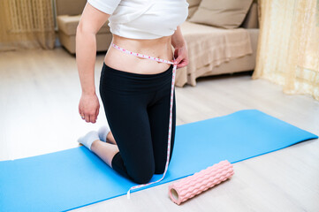 Woman with imperfect waist measuring it with tape while standing on yoga mat, body awareness and...