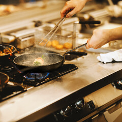 Close-up of chef’s hands preparing food in hot pan on gas stove, with steam rising in a busy restaurant kitchen. Professional cooking process in square format.
