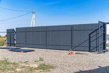 A partially installed corrugated metal fence with an open gate stands on a gravel-covered property,...