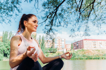 Woman practicing yoga near riverbank, meditating peacefully with city skyline in background,...