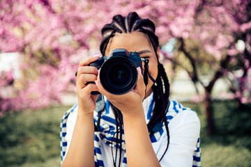 A young woman with braided hair holding a camera in front of cherry blossoms during a sunny spring...
