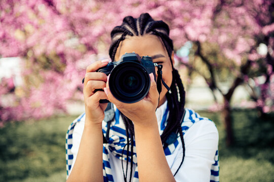 A young woman with braided hair holding a camera in front of cherry blossoms during a sunny spring day outdoors