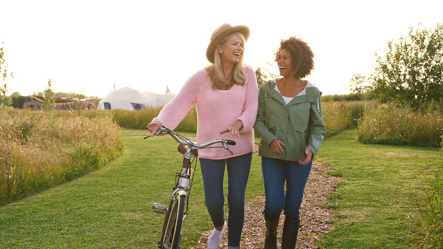 Two Mature Female Friends Walking Along Path With Bike Through Yurt Campsite