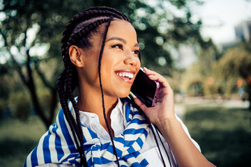 Smiling young woman with braided hair enjoying a phone conversation in a sunny, vibrant urban park...