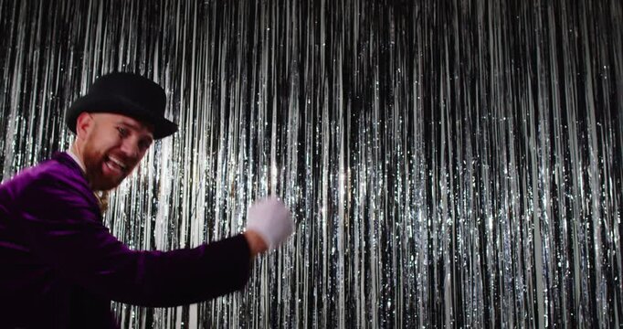 Funny smiling man in magician costume moving backwards and waving his arms against shiny silver curtain. Bearded performer wearing black top hat and purple velvet jacket having fun at disco party.
