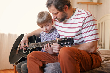 Handsome dad teaches his cute son to play the guitar sitting on sofa in the living room at home....