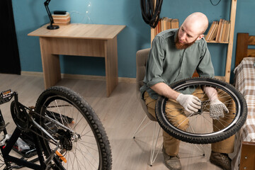 Bearded man mechanic sitting on chair holding bicycle wheel repairing bike at home. Bicycle repair...