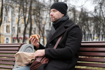 Pensive hipster drinking coffee sitting on a bench in the urban park. Handsome man wearing black...
