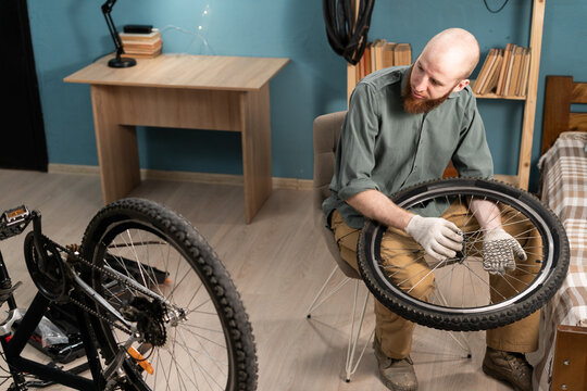 Bearded man mechanic sitting on chair holding bicycle wheel repairing bike at home. Bicycle repair and maintenance