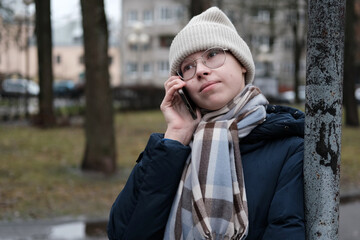 Fototapeta premium Teenager Girl Walking Alone on City Street with Smartphone. Portrait of 14 years old girl wearing eyeglasses, wool hat, winter jacket using mobile phone. Urban Style. Rainy mood weather. Cold season