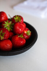 A black bowl filled with fresh, ripe strawberries rests on a clean white table