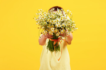 Little boy hiding behind bouquet of beautiful chamomile flowers on yellow background