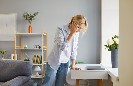 A young blonde woman has a headache, holding her temple with her hand in the apartment. Headache symptom, migraine. Treatment of headache, chronic disease. The problem is brain disease, suffering.