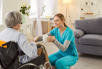 Young friendly female social worker giving a cup of beverage to senior mature woman with disability...