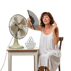 A flushed middle-aged woman fanning herself sits before a table with a vintage fan and a bowl of melting ice cubes, conveying the discomfort of a heatwave or a hot flash, isolated on white background