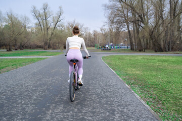 Young woman riding a bicycle in the park on the background of trees. Trendy active girl spends time on the nature. Side view.