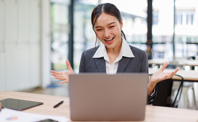 Excited friendly young asian woman 20s in casual clothes sit on desk,  talk work on laptop pc...