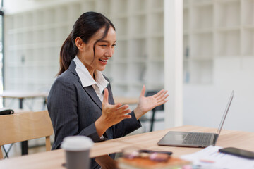 Excited friendly young asian woman 20s in casual clothes sit on desk,  talk work on laptop pc...