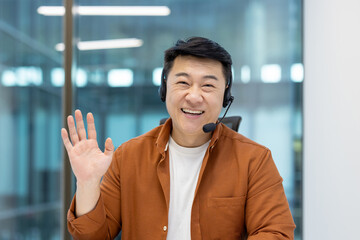 A cheerful Asian man wearing a headset waves at the camera, smiling warmly, in a bright office...