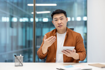 An Asian man in a business setting gestures while holding a tablet, seemingly explaining something.