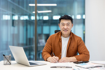 A smiling asian businessman working at his desk, writing and looking at the camera. His office is...
