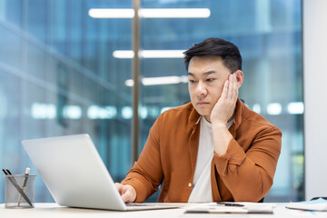 An Asian man looks at a laptop in thought in a modern office setting, contemplating and making...