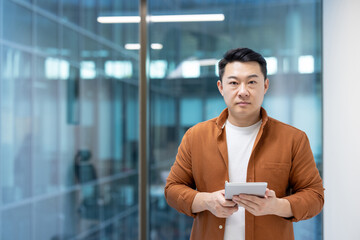 A focused Asian man holds a tablet, posing indoors in a modern office environment, looking directly...